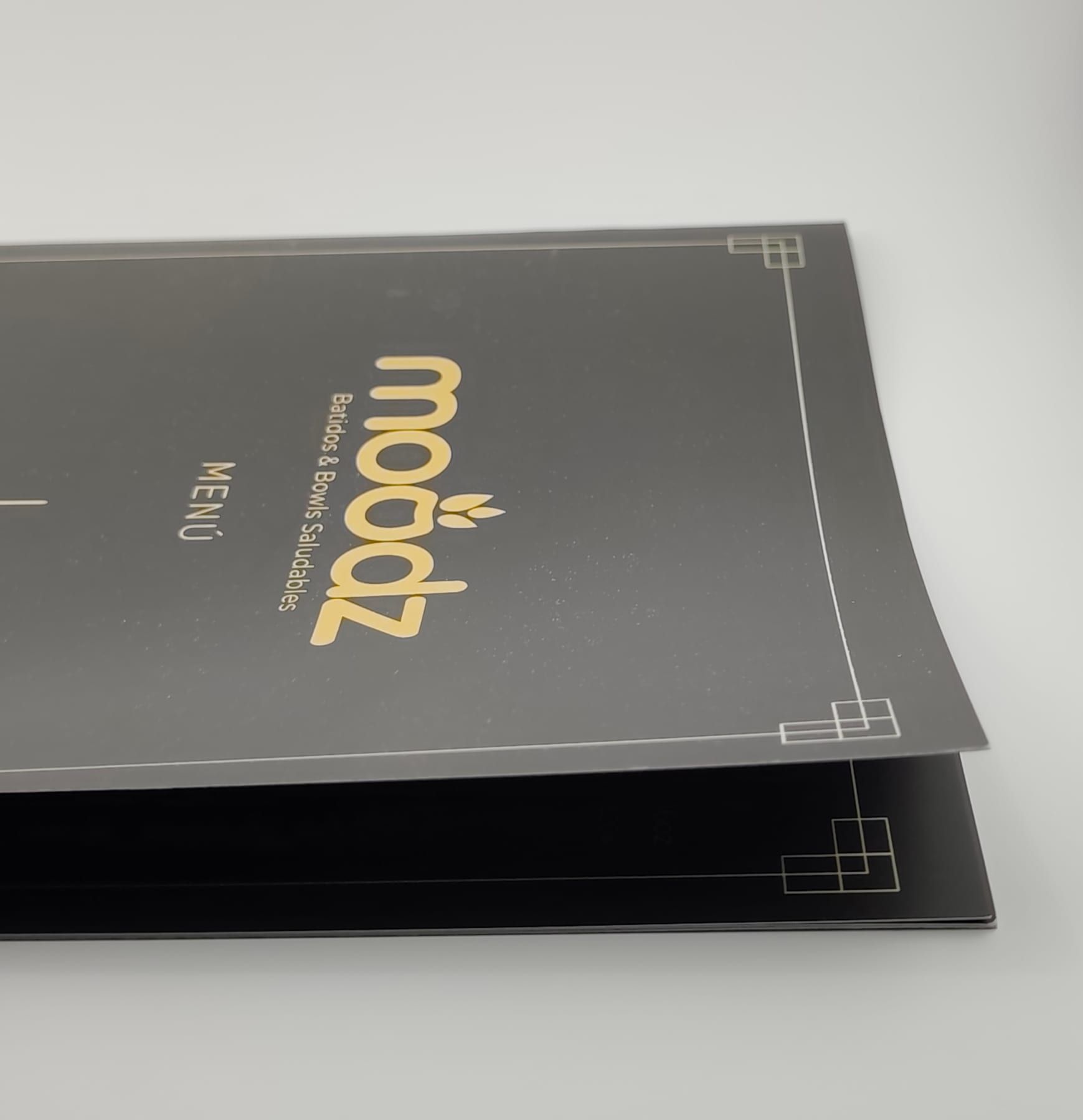 Carpeta menú en negro mate con logotipo estampado en dorado y filetes plateados, mostrando el plegado del lomo