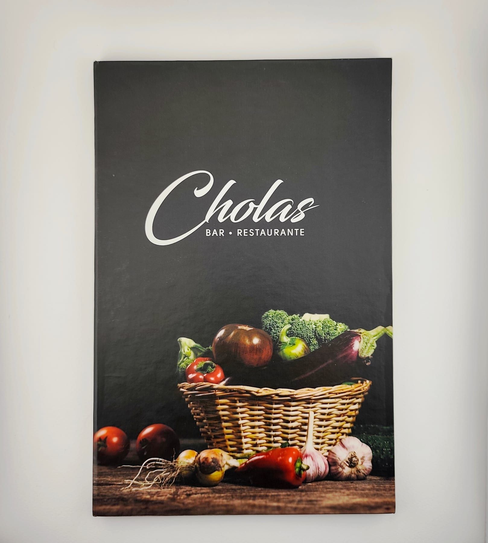 Carta de tapa dura del restaurante Cholas cerrada, con portada negra impresa a todo color con fotografía de vegetales