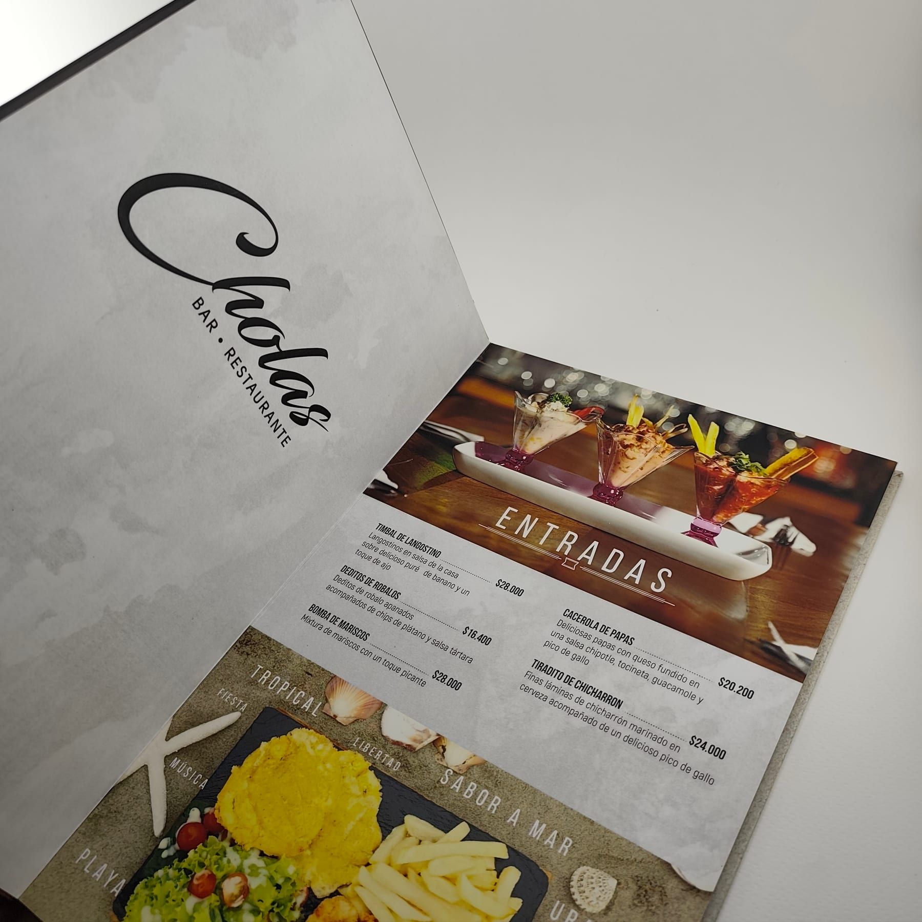 Menú de pasta dura para restaurante abierto, mostrando la guarda con el logo de Cholas y una página interior de entradas con fotografía gastronómica a todo color
